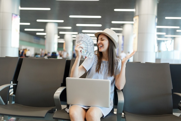 Young joyful traveler tourist woman working on laptop, holds bundle of dollars, cash money spread hands wait in lobby hall at airport