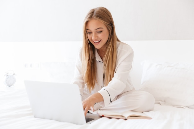 young joyful woman in pajama typing on laptop and smiling while sitting on bed in light room