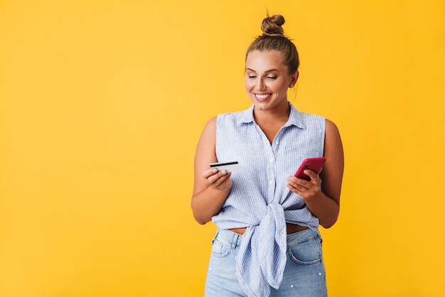 Young joyful woman in shirt happily holding credit card and cellphone in hands over yellow background isolated