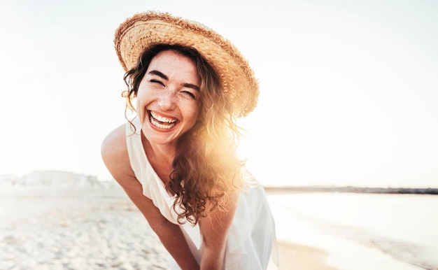 Young joyful woman in white shirt wearing hat smiling at camera on the beach