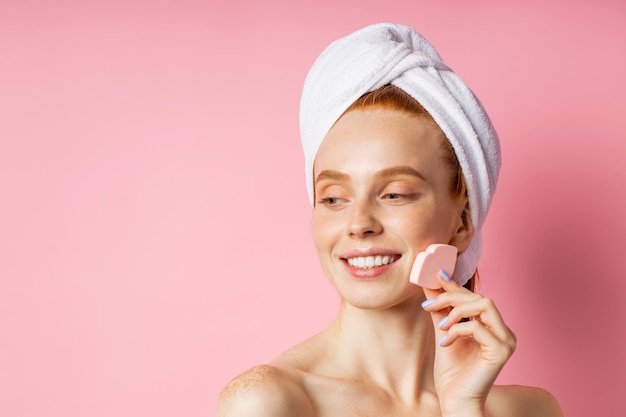 Young joyful woman with white towel on head, fresh pure skin keeping cosmetic sponge near face, smiling looking aside on pink background with copy space for text, advertising. Beauty products.