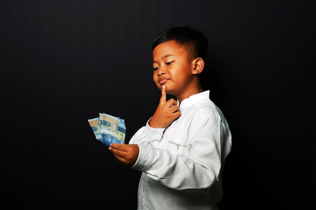 Young kid with confused expression while holding a lot of money isolated on black background