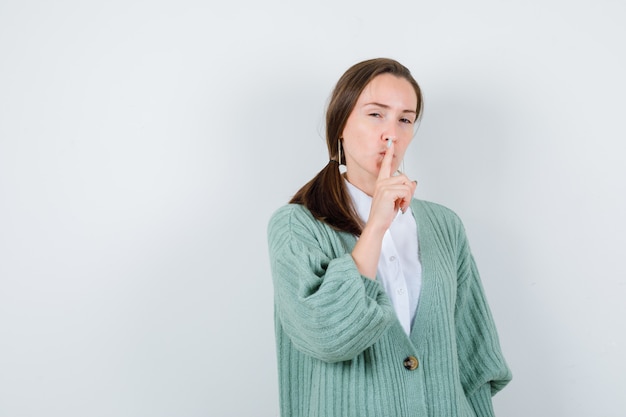 Young lady in blouse, cardigan showing silence gesture and looking sensible , front view.