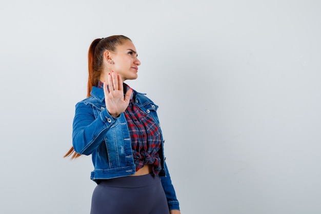 Young lady in checkered shirt, denim jacket showing stop gesture and looking confident , front view.