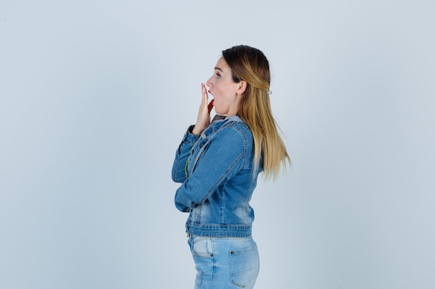 Young lady keeping hand on mouth in denim outfit and looking shocked. .