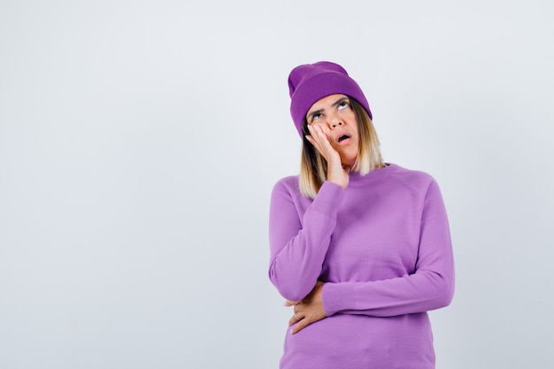 Young lady leaning cheek on palm in purple sweater, beanie and looking perplexed. front view.