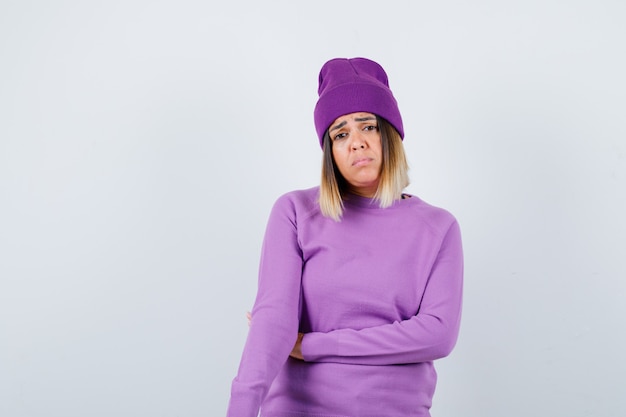 Young lady looking at camera while frowning face in purple sweater, beanie and looking disappointed. front view.