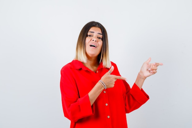 Young lady pointing aside in red oversize shirt and looking excited. front view.