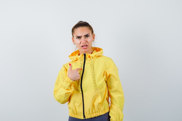 Young lady pointing at herself as asking question in yellow jacket and looking serious , front view.