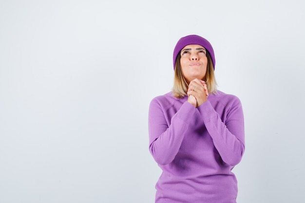 Young lady in purple sweater, beanie holding clasped hands in praying gesture and looking hopeful , front view.