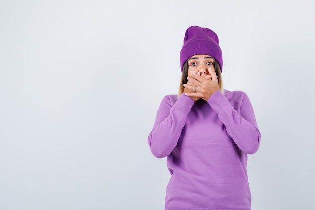 Young lady in purple sweater, beanie with hands on mouth and looking scared , front view.