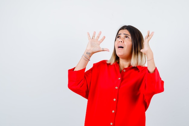 Young lady in red oversize shirt keeping hands in surrender gesture and looking anxious , front view.