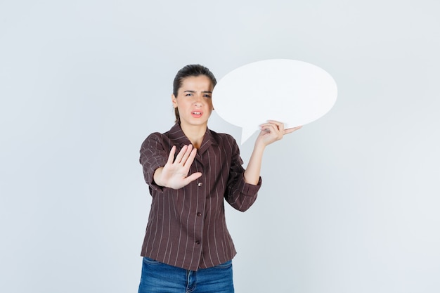 Young lady in shirt, jeans showing stop gesture, keeping paper poster and looking disappointed , front view.