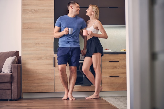  young lady in shorts and brunette man spending time in cosy atmosphere while drinking tea