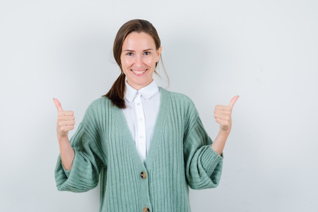 Young lady showing double thumbs up in blouse, cardigan and looking joyful. front view.