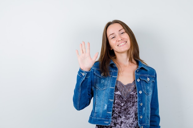 Young lady waving hand for greeting in blouse, denim jacket and looking cheerful , front view.