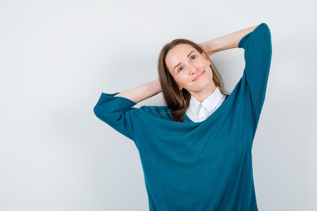 Young lady in white shirt, sweater looking away with hands behind head and looking dreamy , front view.