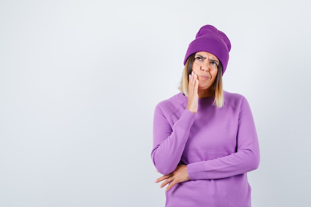 Young lady with hand on cheek in purple sweater, beanie and looking pensive. front view.