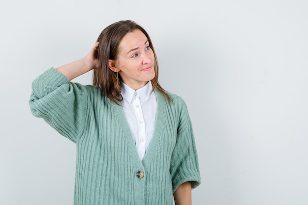 Young lady with hand behind head in shirt, cardigan and looking pleased. front view.