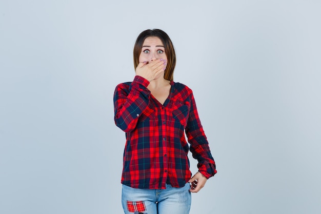 Young lady with hand on mouth in checked shirt, jeans and looking terrified. front view.