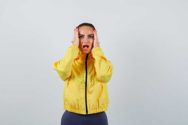 Young lady with hands on head, opening mouth in yellow jacket and looking terrified . front view.