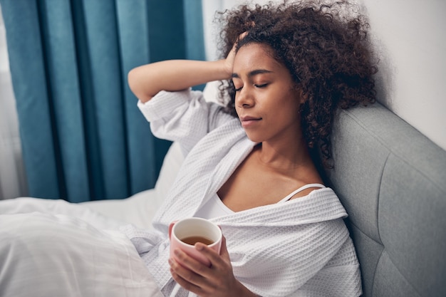 Young lady with a mug of green tea in her hand dozing in her bedroom