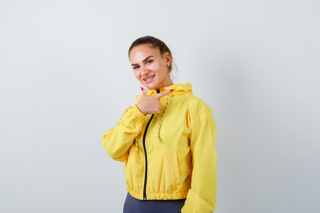 Young lady in yellow jacket pointing to the right side and looking merry , front view.