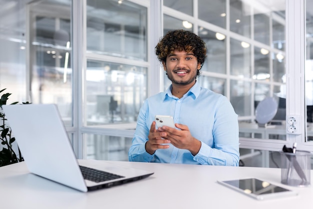 A young latin american businessman sits in the office at a desk and uses a mobile phone writes a