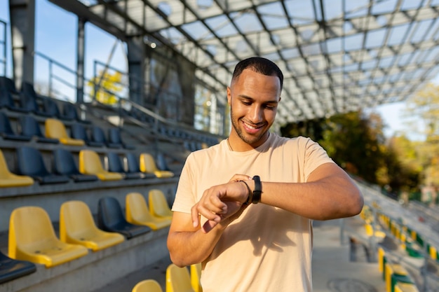 Young latin american male sportsman stands at the stadium and looks at the fitness bracelet the