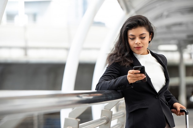 Young Latin businesswoman using cell phone and holding handle of trolley bag 