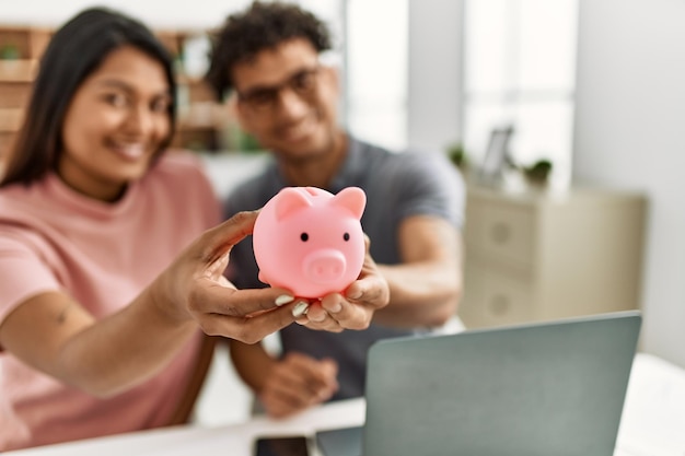 Young latin couple smiling happy holding piggy bank sitting on the table at home