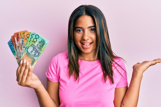 Young latin girl holding australian dollars celebrating achievement with happy smile and winner expression with raised hand