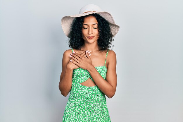 Young latin girl wearing summer hat smiling with hands on chest with closed eyes and grateful gesture on face. health concept.