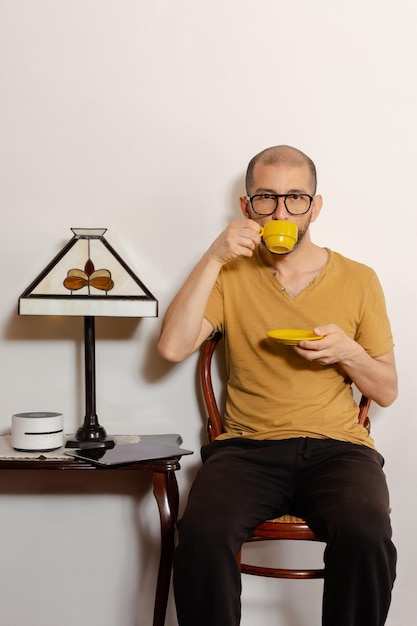 Young latin man in casual clothing and glasses drinking coffee sitting on a chair at home Copy space