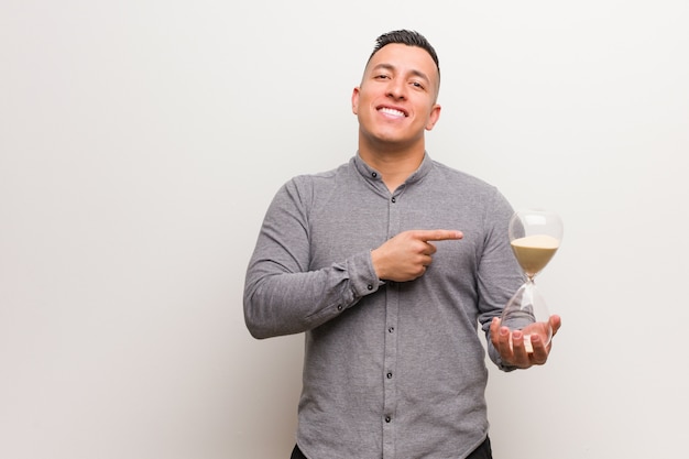 Young latin man holding a sand timer pointing to the side with finger