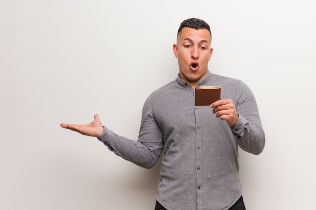 Young latin man holding a wallet holding something on palm hand