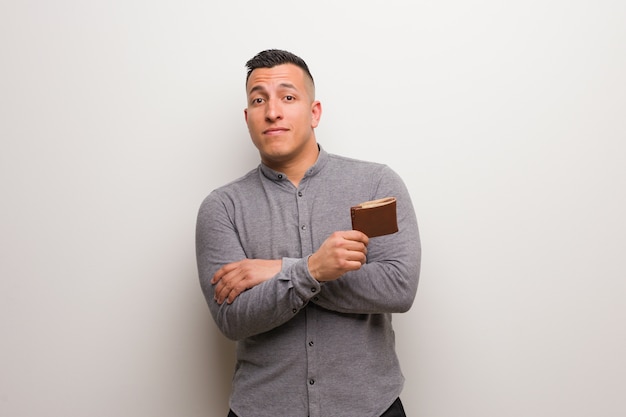 Young latin man holding a wallet smiling confident and crossing arms, looking up