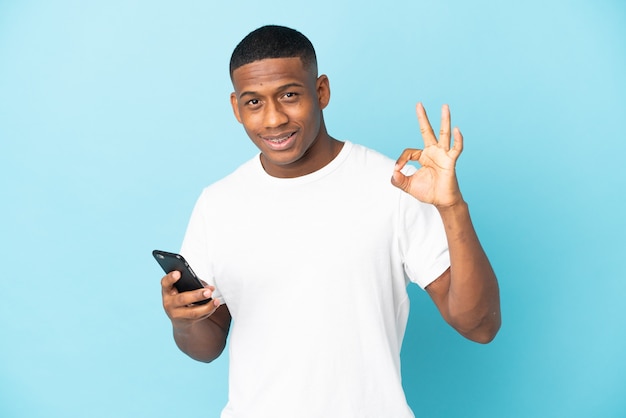 Young latin man isolated on blue wall using mobile phone and doing OK sign