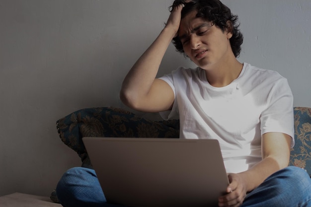 Young latin man with his hand on his head stressing out looking at his laptop screen while working in his bedroom sitting on his bed