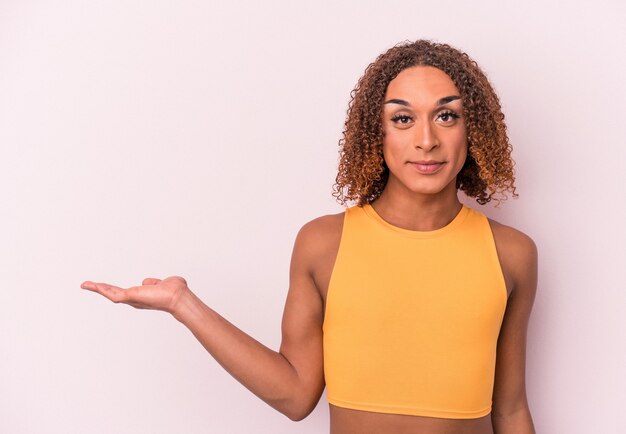 Young latin transsexual woman isolated on pink background showing a copy space on a palm and holding another hand on waist.