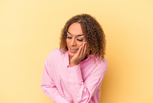 Young latin transsexual woman isolated on yellow background laughs happily and has fun keeping hands on stomach.