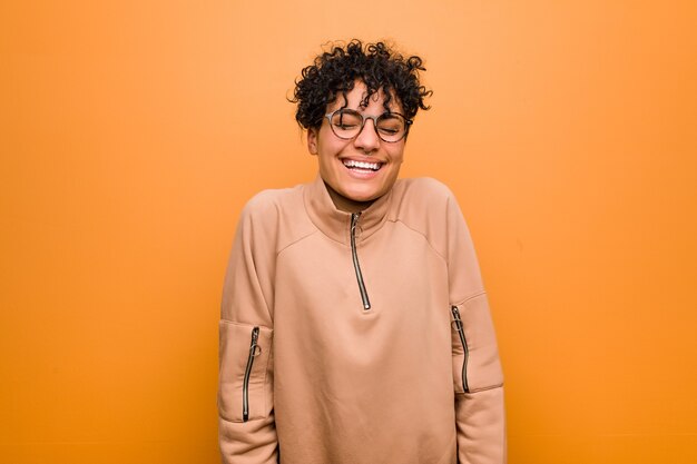 Young latin woman against a brown wall laughs and closes eyes, feels relaxed and happy.