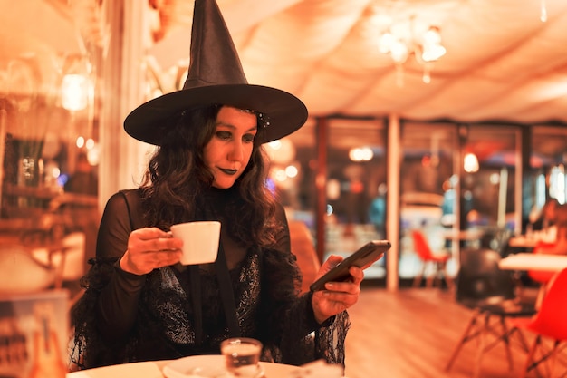 Young latin woman dressed as a witch sitting having a coffee and looking at the cell phone in a cafe