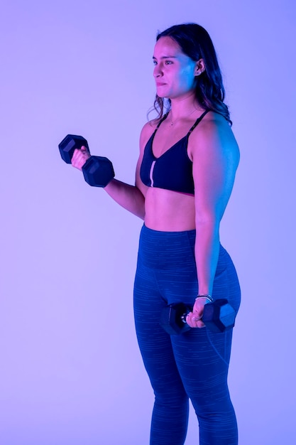 Young latin woman exercising with dumbbells standing in studio with color background