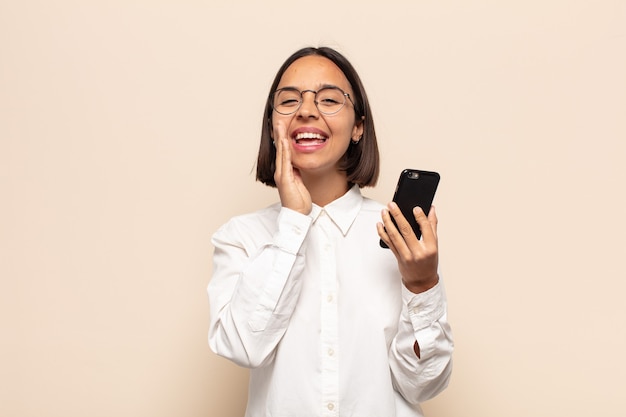 Young latin woman feeling happy, excited and positive, giving a big shout out with hands next to mouth, calling out