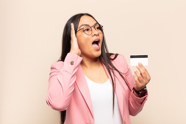 Young latin woman feeling happy, excited and surprised, looking to the side with both hands on face