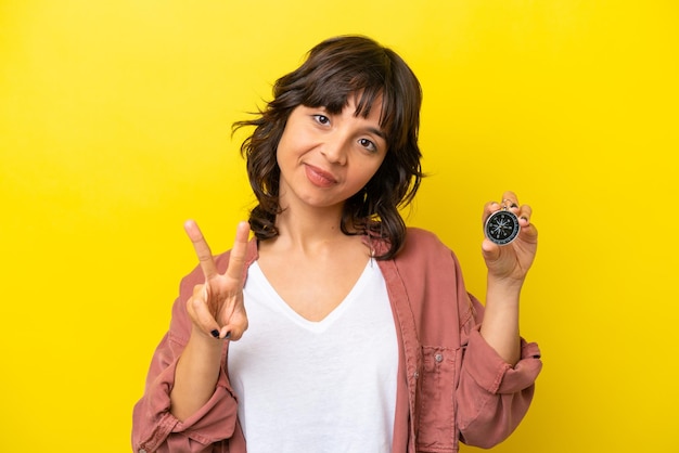 Young latin woman holding compass isolated on yellow background smiling and showing victory sign