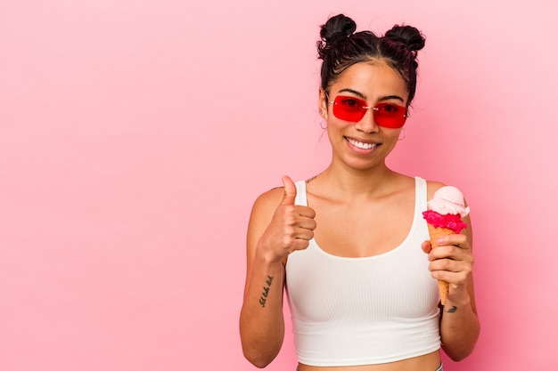 Young latin woman holding an ice cream isolated on pink background smiling and raising thumb up