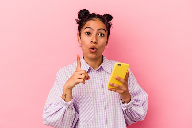 Young latin woman holding a mobile phone isolated on pink background having some great idea, concept of creativity.