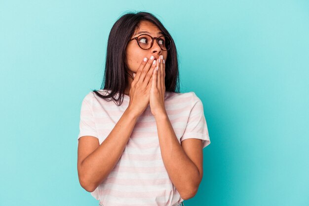 Young latin woman isolated on blue background thoughtful looking to a copy space covering mouth with hand.